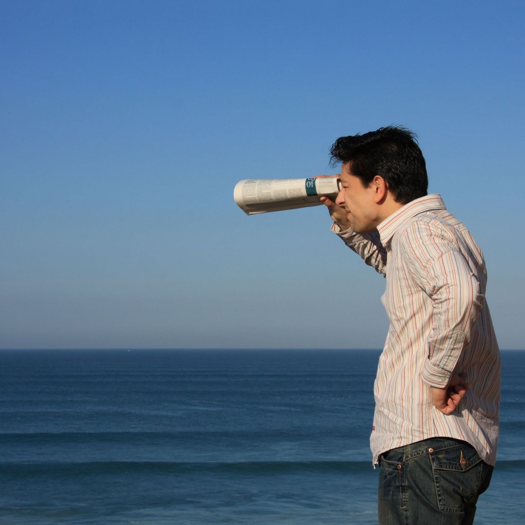 photo: a man in a striped shirt looks through a rolled-up newspaper toward the ocean under a blue sky.